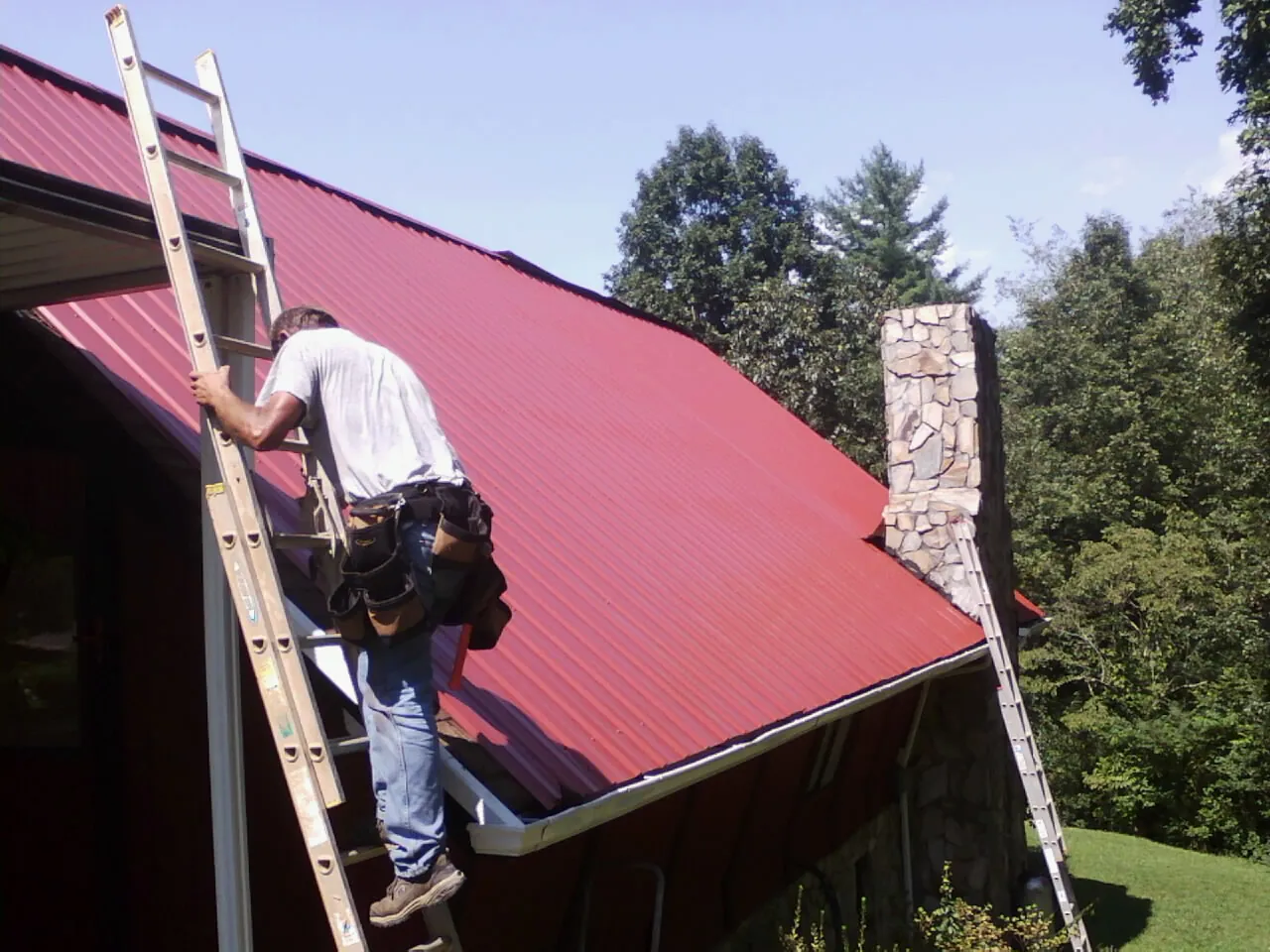 Roofer on a ladder beside a red metal roof and stone chimney — Soffit & Fascia Repair by Roofing Brothers in Allen Park